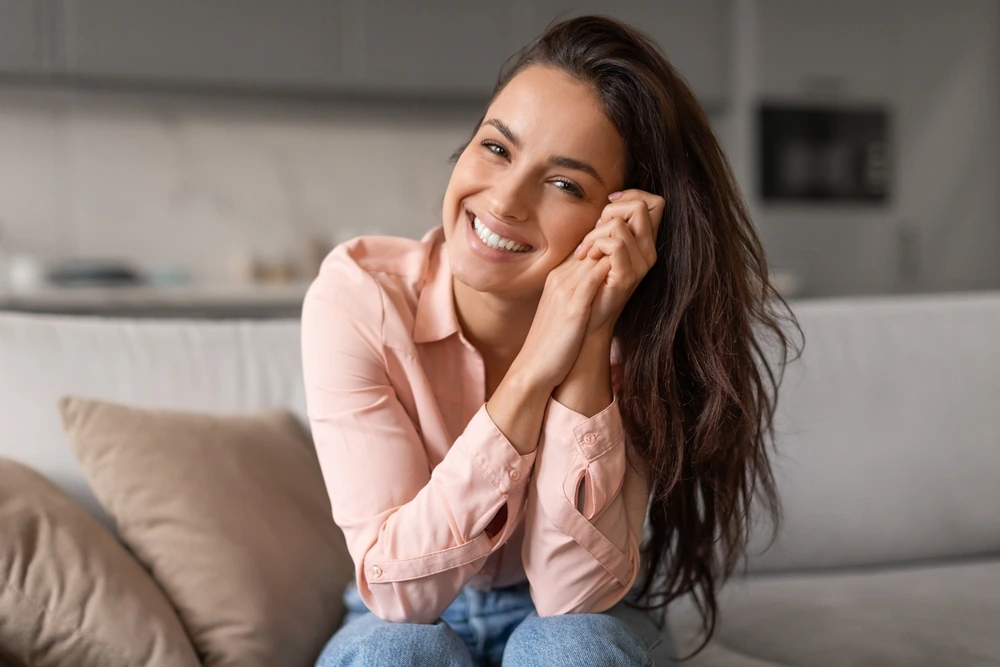 Radiant,Woman,With,Contagious,Smile,Poses,Playfully,,Hands,Gently,Cradling A smiling woman seated on a sofa